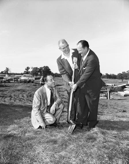 two men, wearing suts, hold a shovel in the ground. another man in a suit is kneeling next to the shovel. All 3 men have a smiles on their faces and looking at each other