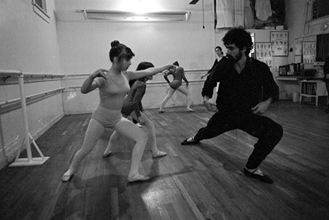 A man is teaching a girl, wearing a leotard, some dance steps. There are other girls in leotards standing behind them.