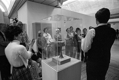 A group of people stand around a glass display case. A woman is describing the object in the case while a man signs what she is 