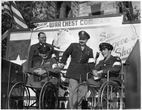men in uniform, two of whom are in wheelchairs, pose in front of poster "Texas War Chest Command"