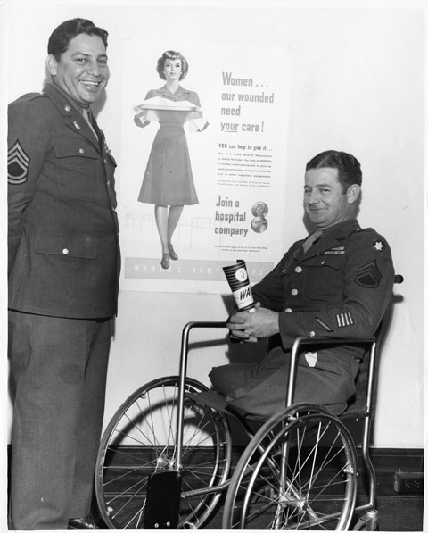 Two men in military uniform, one in a wheelchair, pose beside poster advertising to women to join a hospital company