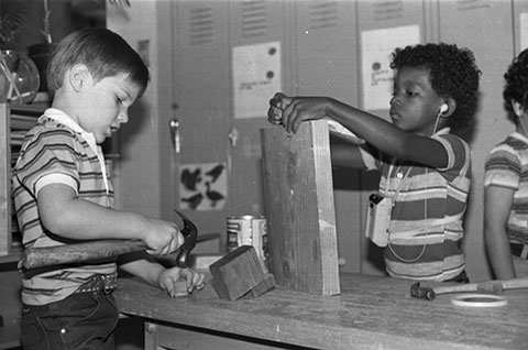 Two boys are working at a table. One is hammering a nail into a block of wood. The other is holding a block of wood.