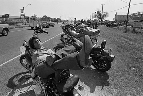 Joan Bolten rests on motorcycle. several motorcycles are parked and lined up along the road behind him.