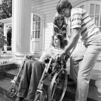 a man and woman help a woman, who is in a wheelchair, up some stairs