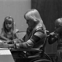 woman in wheelchair is at a table folding paper; two girls are standing beside her