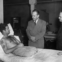 Shirtless man with both legs amputated in a hospital bed being visited by two men in suits