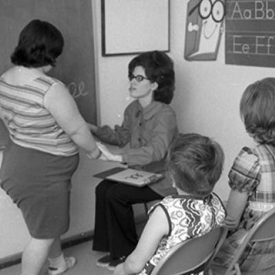 woman sitting in chair helping student standing in front of her,  three children sitting in chairs