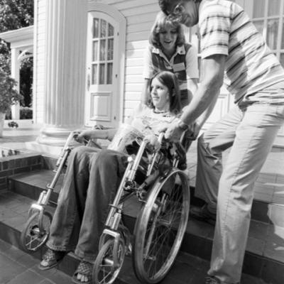 a man and woman help a woman, who is in a wheelchair, up some stairs