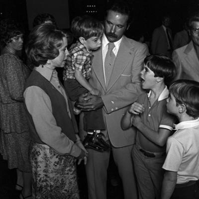 A boy is being held by a man wearing a suit. There are two boys standing next to the man. They are talking to a woman.
