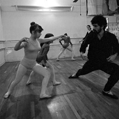 A man is teaching a girl, wearing a leotard, some dance steps. There are other girls in leotards standing behind them.