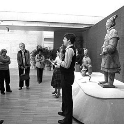 A man and woman stand in front a group of people. Behind the man and woman are statues.