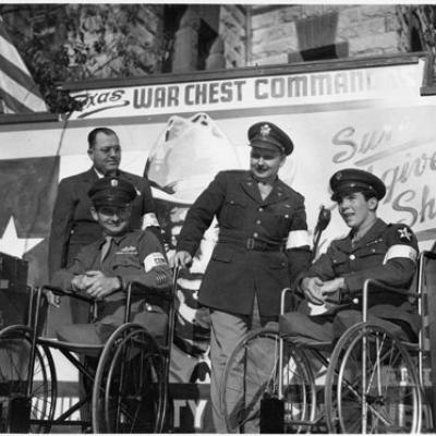 men in uniform, two of whom are in wheelchairs, pose in front of poster "Texas War Chest Command"