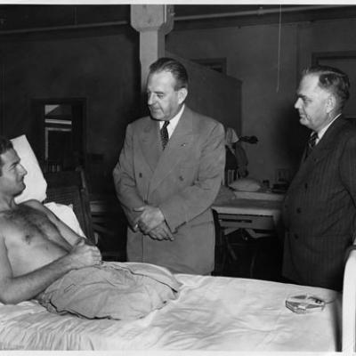 Shirtless man with both legs amputated in a hospital bed being visited by two men in suits