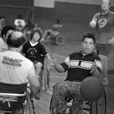 4 men playing wheelchair soccer, a man with a whistle is standing in the background